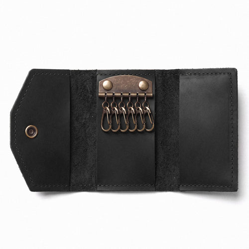 A black leather key ring case shown open on a white background, featuring a brass-toned metal key ring holder with six key rings and snap-button closure.