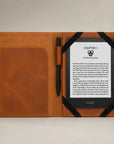 A tan leather folio-style Kindle case shown open on a light neutral background, with a black pen in a side pocket and a Kindle e-reader partially inserted on the right side.