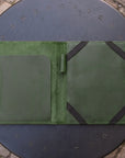 An open green leather folio or organizer laid flat on a round black metal table outdoors. The folio has a suede-like center panel, a large smooth pocket on the left, and two black elastic bands attached to the right side.