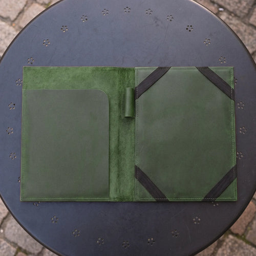 An open green leather folio or organizer laid flat on a round black metal table outdoors. The folio has a suede-like center panel, a large smooth pocket on the left, and two black elastic bands attached to the right side.