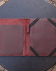 A dark reddish-brown leather folio or organizer spread open on a round black metal table outdoors. The left side has a large smooth pocket, and the right side has a textured panel secured with black elastic bands.