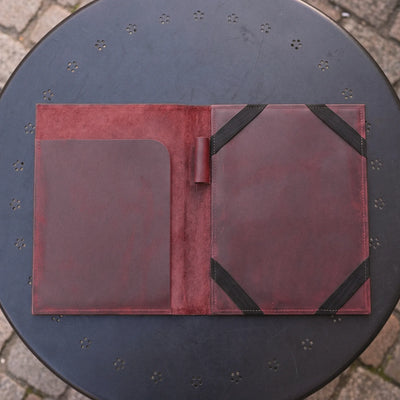 A dark reddish-brown leather folio or organizer spread open on a round black metal table outdoors. The left side has a large smooth pocket, and the right side has a textured panel secured with black elastic bands.
