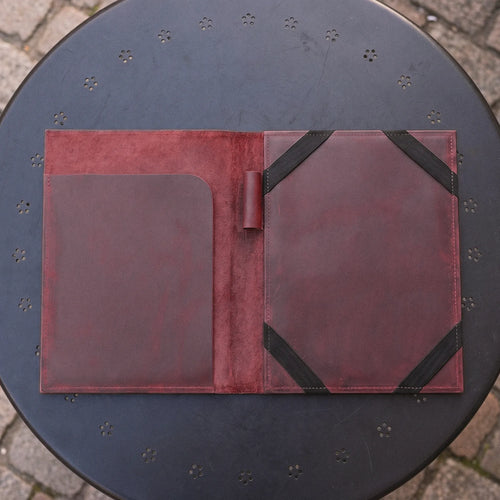 A dark reddish-brown leather folio or organizer spread open on a round black metal table outdoors. The left side has a large smooth pocket, and the right side has a textured panel secured with black elastic bands.