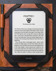 A Kindle e-reader device in a brown leather folio case, displaying a book chapter on a dark textured surface.