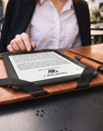 A person seated at an outdoor cafe uses a black e-reader with a tan leather folio case and a matching stylus. The e-reader screen shows a book page with the visible brand/title text 'CHAPTER 1' and 'THE STORY OF THE SAID.