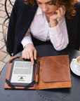 A woman in a navy blazer and white shirt sits at a round black table outdoors, using a brown leather e-reader case with a black strap holding an e-reader and a pen. A cup of coffee is on the table beside her.
