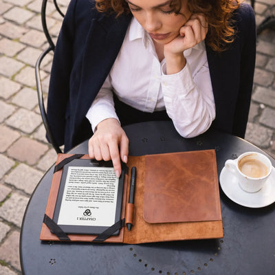 A woman in a navy blazer and white shirt sits at a round black table outdoors, using a brown leather e-reader case with a black strap holding an e-reader and a pen. A cup of coffee is on the table beside her.