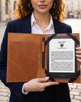A woman in a navy blazer and white shirt holds an open brown leather folio case containing a black Kindle e-reader with a black cover and a black pen, set against a blurred cobblestone street background.