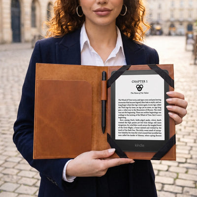 A woman in a navy blazer and white shirt holds an open brown leather folio case containing a black Kindle e-reader with a black cover and a black pen, set against a blurred cobblestone street background.