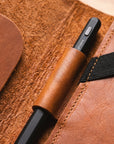 A close-up of a black pen with a brown leather sleeve, resting inside a brown leather organizer pocket with a black elastic band.