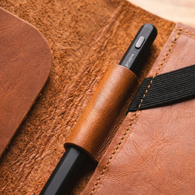A close-up of a black pen with a brown leather sleeve, resting inside a brown leather organizer pocket with a black elastic band.