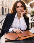 A woman in a white shirt and navy blazer sits at an outdoor cafe table with a brown leather folder in front of her. The background shows a blurred cobblestone street and cafe seating.