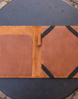 A brown leather organizer sleeve or folio laid open on a round black metal table outdoors. The left side has a smooth brown pocket, and the right side has a larger panel with black elastic bands holding small items.