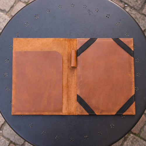 A brown leather organizer sleeve or folio laid open on a round black metal table outdoors. The left side has a smooth brown pocket, and the right side has a larger panel with black elastic bands holding small items.
