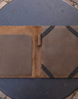 A brown leather portfolio folder or notebook cover shown open, placed on a round dark metal outdoor table with a stone-paved ground visible around it.