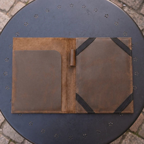 A brown leather portfolio folder or notebook cover shown open, placed on a round dark metal outdoor table with a stone-paved ground visible around it.