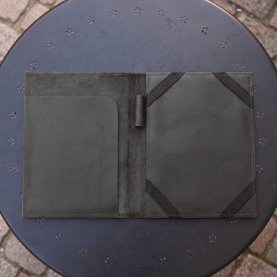 A dark olive green leather portfolio folder shown open, resting on a round black metal patio table with a stone paver floor visible in the background.