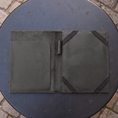 A dark olive green leather portfolio folder shown open, resting on a round black metal patio table with a stone paver floor visible in the background.