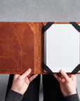 A person holds open a brown leather tablet folio case, showing a white tablet inside with black corner straps, against a gray textured background.