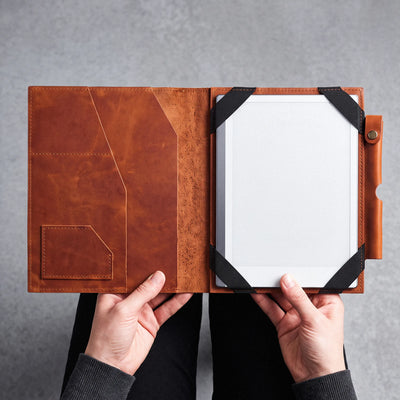 A person holds open a brown leather tablet folio case, showing a white tablet inside with black corner straps, against a gray textured background.