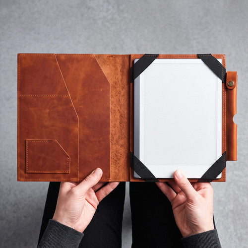 A person holds open a brown leather tablet folio case, showing a white tablet inside with black corner straps, against a gray textured background.
