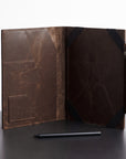 A dark brown distressed leather folio case shown open, displaying its interior pockets and texture, with a black stylus resting in front on a dark surface against a white background.