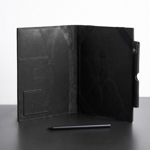 A black leather folio-style tablet sleeve or organizer, shown open on a dark surface against a light gray background. The sleeve has a textured finish, a pocket on the left panel, and a pen-like stylus placed in front.