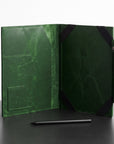 A dark green leather folio or tablet sleeve shown open, with black corner protectors and a pen resting in front on a dark surface against a light background.