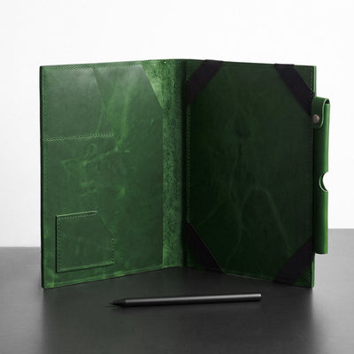 A dark green leather folio or tablet sleeve shown open, with black corner protectors and a pen resting in front on a dark surface against a light background.