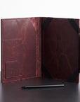 A dark reddish-brown leather folio case shown open on a dark tabletop against a light gray background, with black corner straps and a small black stylus placed in front.