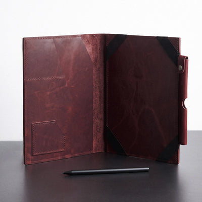 A dark reddish-brown leather folio case shown open on a dark tabletop against a light gray background, with black corner straps and a small black stylus placed in front.