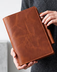 A person holding a brown leather planner or binder cover with a snap-button closure. The hands and torso of the person are visible, wearing a dark gray knit sweater against a light gray background.