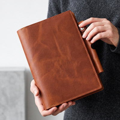 A person holding a brown leather planner or binder cover with a snap-button closure. The hands and torso of the person are visible, wearing a dark gray knit sweater against a light gray background.