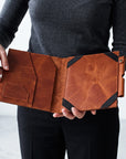 A person holding an open brown leather folio or tablet sleeve with black corner accents, shown against a plain light gray background.