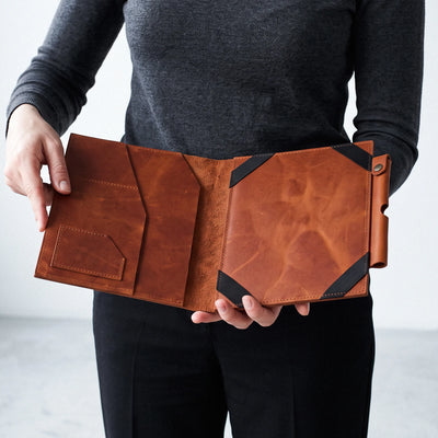A person holding an open brown leather folio or tablet sleeve with black corner accents, shown against a plain light gray background.