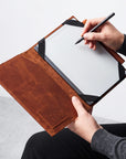 A person holds an open brown leather folio-style tablet case while writing on a tablet with a black stylus. The case has black corner straps and a visible interior pocket, shown against a light neutral background.