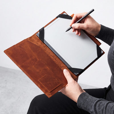 A person holds an open brown leather folio-style tablet case while writing on a tablet with a black stylus. The case has black corner straps and a visible interior pocket, shown against a light neutral background.