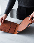 A person holding open a brown leather folio or document holder on a light gray tabletop. The product shows warm tan and medium brown leather panels with black corner accents and a small pocket on the inside flap.