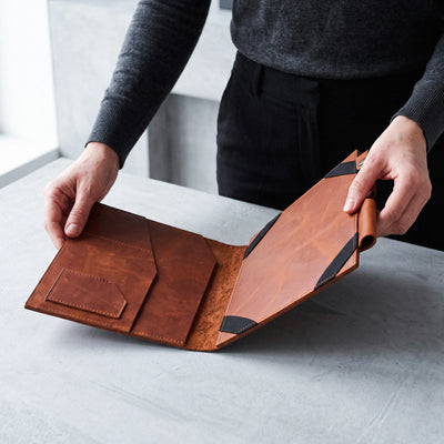 A person holding open a brown leather folio or document holder on a light gray tabletop. The product shows warm tan and medium brown leather panels with black corner accents and a small pocket on the inside flap.