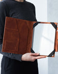 A person in a dark gray long-sleeve shirt holds an open brown leather tablet sleeve or document holder with black corner straps and a clear front panel.
