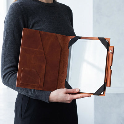A person in a dark gray long-sleeve shirt holds an open brown leather tablet sleeve or document holder with black corner straps and a clear front panel.