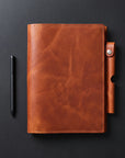 A brown leather planner cover with a snap-button strap closure, shown flat on a dark gray background next to a black pen.