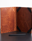 A brown leather folio or tablet sleeve shown open on a dark tabletop against a light background, with black corner straps and a small pen placed in front.