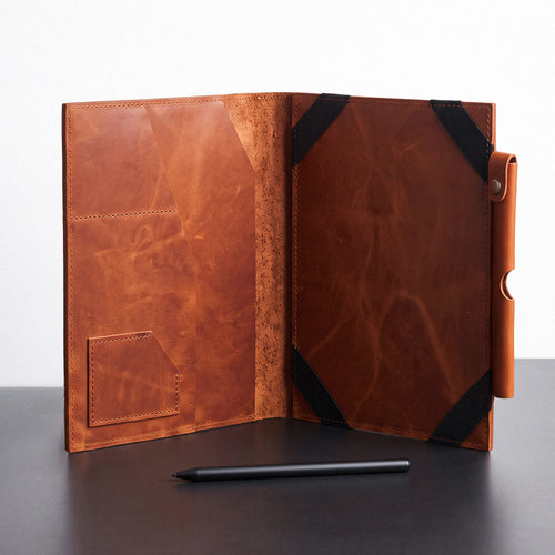 A brown leather folio or tablet sleeve shown open on a dark tabletop against a light background, with black corner straps and a small pen placed in front.