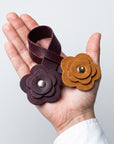 Two leather flower-shaped wristlets or bracelets held in a hand against a light gray background. One is a deep burgundy/brown color with a silver snap, and the other is a warm tan/caramel brown with a gold snap.