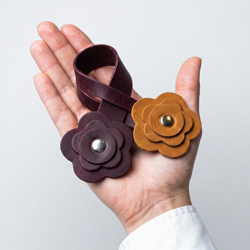 Two leather flower-shaped wristlets or bracelets held in a hand against a light gray background. One is a deep burgundy/brown color with a silver snap, and the other is a warm tan/caramel brown with a gold snap.