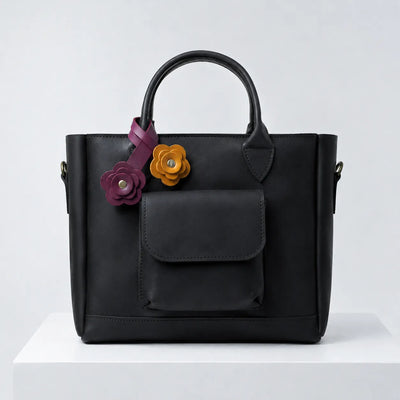 A black leather handbag with dual top handles, a front flap pocket, and two decorative flower charms in purple and mustard yellow, displayed on a white pedestal against a light gray background.