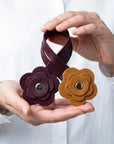 A person in a white shirt holds a dark burgundy leather loop with two flower-shaped metal snap closures, one dark burgundy and one mustard yellow, against a white background.