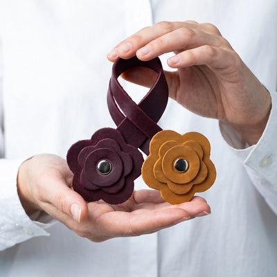 A person in a white shirt holds a dark burgundy leather loop with two flower-shaped metal snap closures, one dark burgundy and one mustard yellow, against a white background.