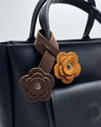 A close-up of a black leather handbag with two decorative flower-shaped leather charms attached to the handle. One charm is medium brown and the other is mustard yellow, both with metal snap buttons.
